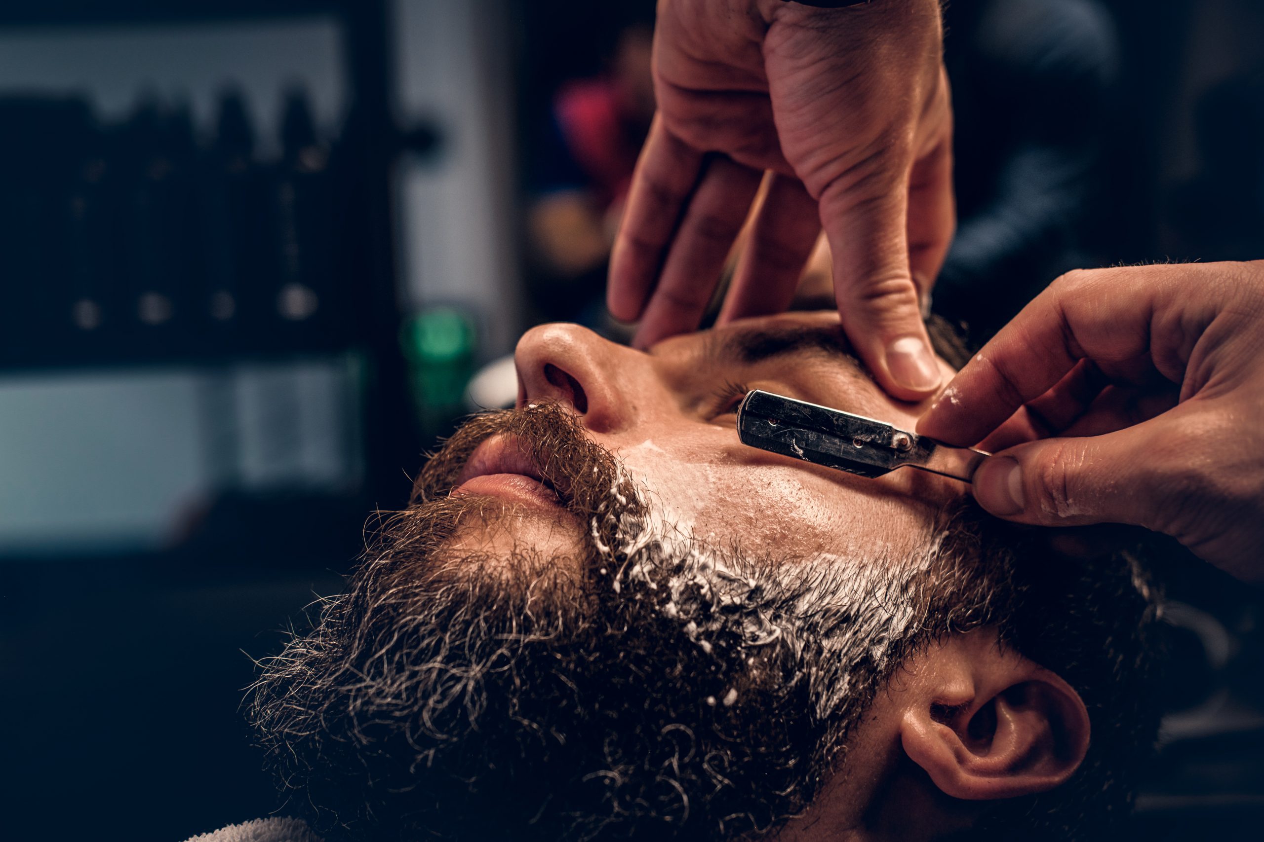 Close up portrait of barber shaving bearded male with a sharp razor in a saloon.
